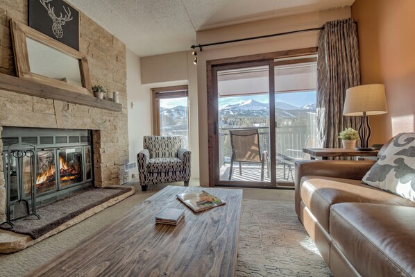 A cozy living room with a fireplace, a leather sofa, a patterned armchair, and a view of mountains through a large sliding glass door. - Village at Breckenridge - Shavano Unit 2603 Ski In/Out (Breckenridge)