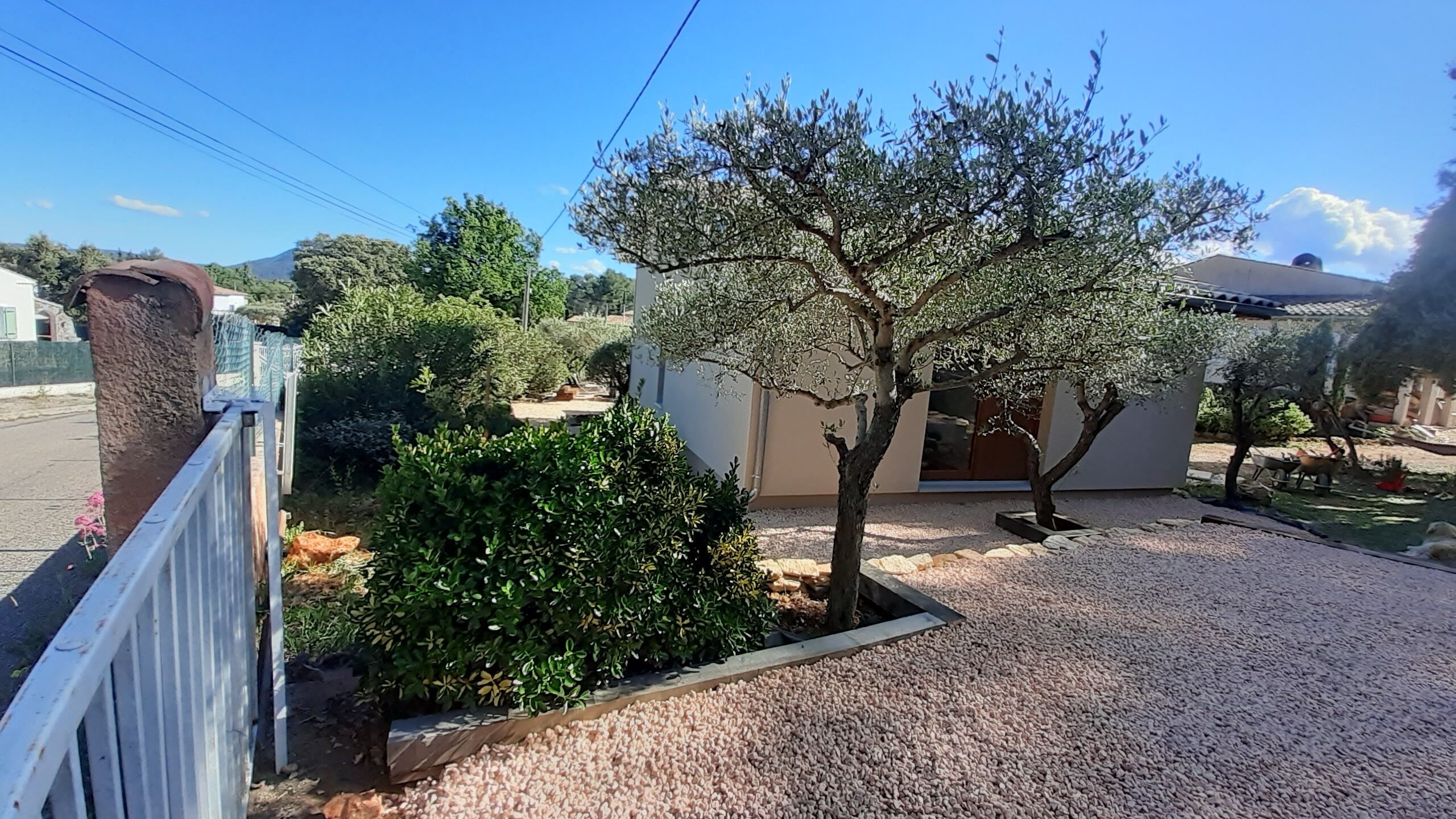A residential property with a gravel driveway, a white fence, and a tree in the foreground.