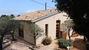 Une maison de plain-pied avec un toit en tuiles, une allée gravillonnée et un petit jardin agrémenté d’oliviers et d’arbustes.