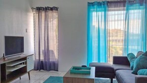 A living room with a flat-screen TV, a sofa, a coffee table, and a view of the outdoors through sheer curtains.