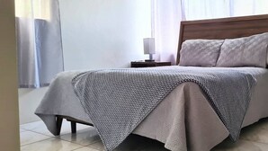 A neatly made bed with a patterned bedspread and pillows, a bedside table with a lamp, and sheer curtains allowing natural light.