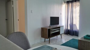 A room with a television on a wooden stand, a door, a window with curtains, and a table with a blue rug.