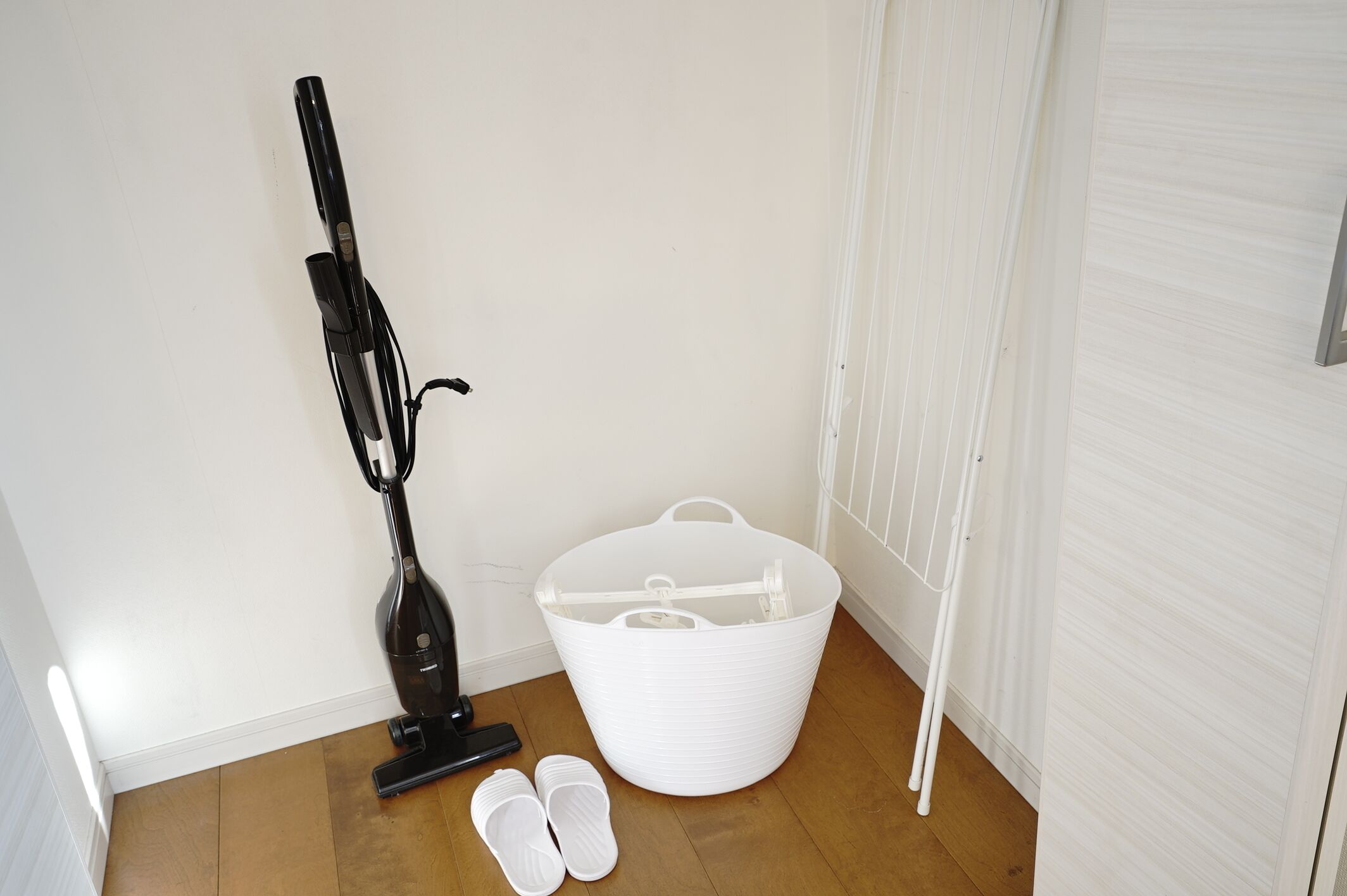 A corner of a room with a white plastic storage bin, a black vacuum cleaner, and a pair of white slippers on the floor.