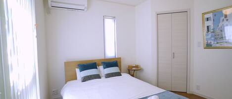 A hotel room with a bed, a bedside table, an air conditioning unit, a window, a door, and a painting on the wall.