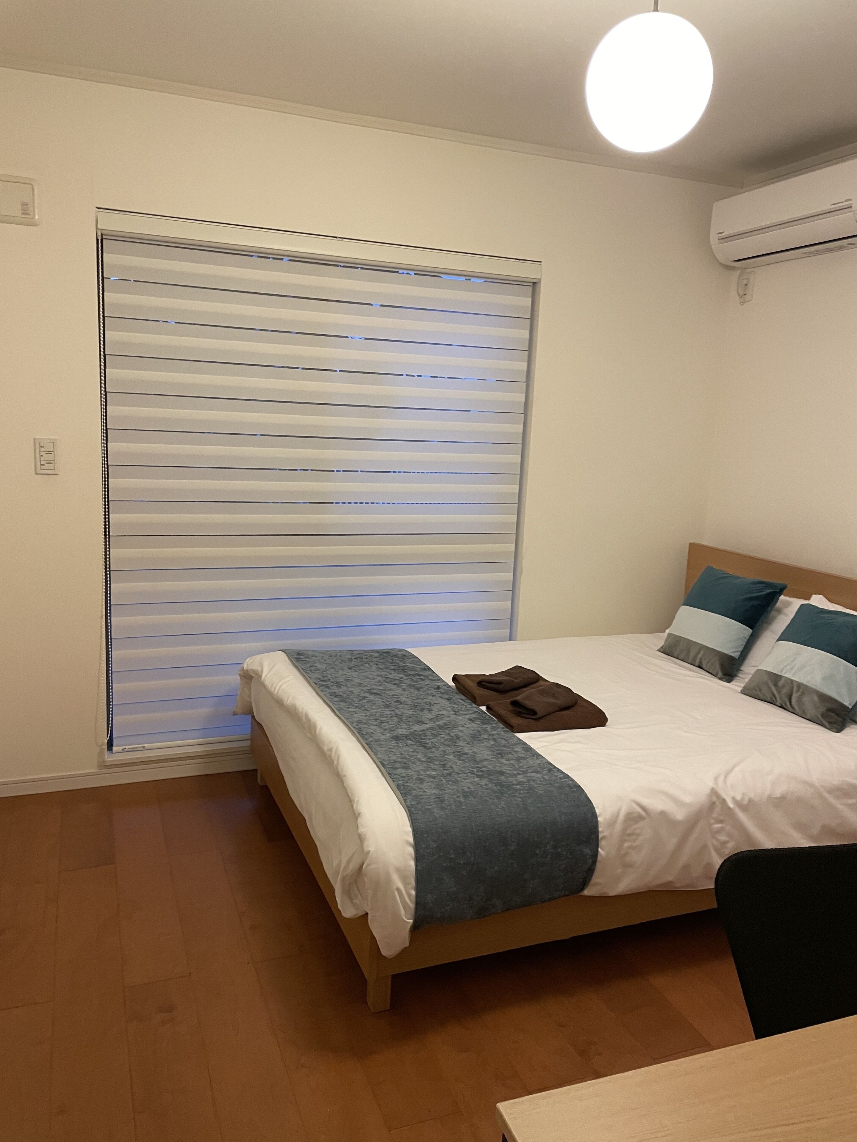 Ein Schlafzimmer mit Bett, Nachttisch, Fenster mit Jalousien und Klimaanlage.