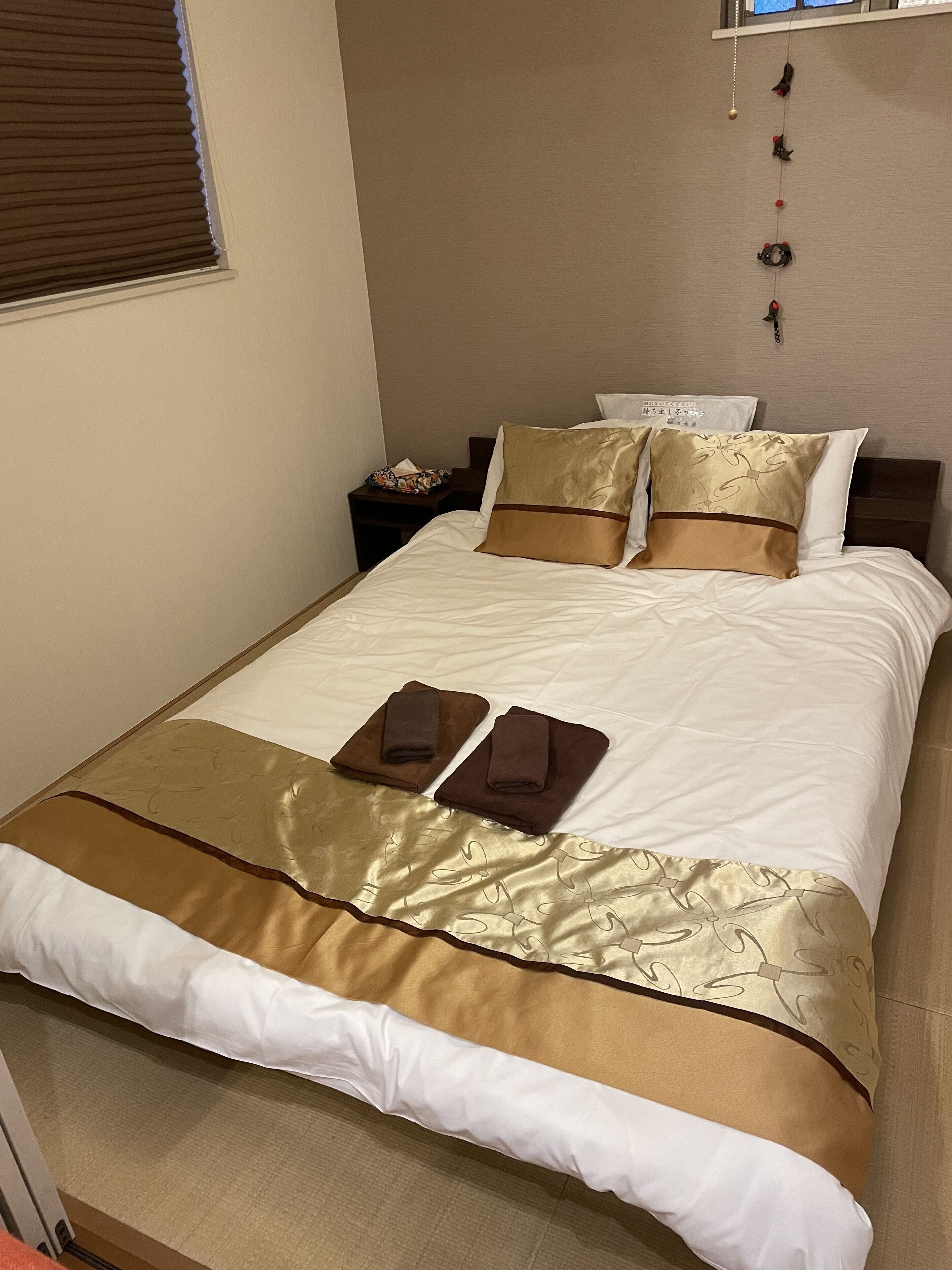 Una cama bien hecha con una colcha dorada y blanca, dos toallas dobladas y una mesita de noche con una lámpara y una caja de pañuelos.