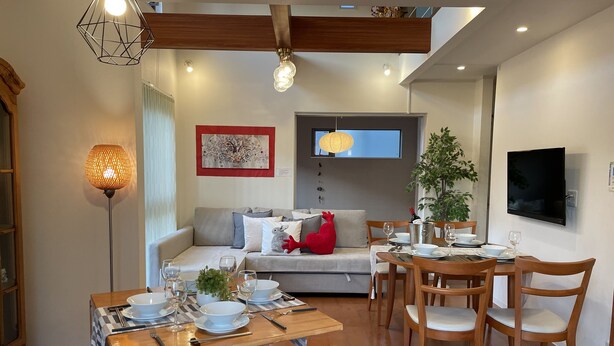 A modern living room with a dining area, wooden furniture, a sofa with cushions, a red heart-shaped pillow, a wall-mounted TV, and a floor lamp. - Skytree House In Tokyo (Tokyo)