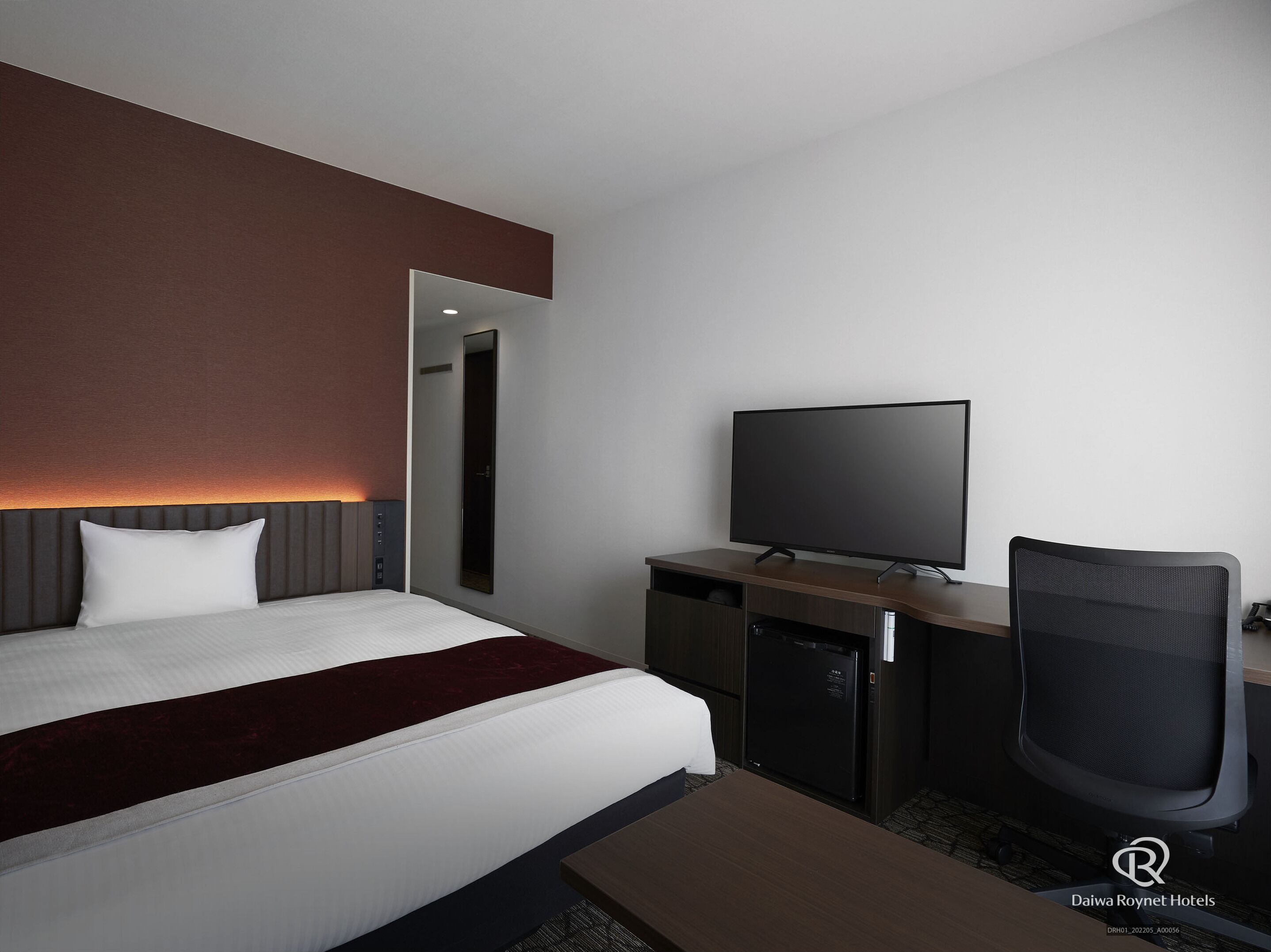 A modern hotel room with a bed, a desk with a flat-screen TV, a chair, and a minibar.