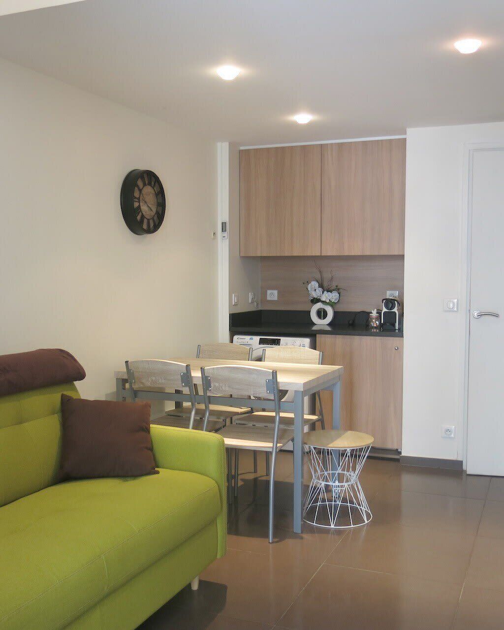 A compact living space with a green sofa, a dining table with chairs, a kitchen area with wooden cabinets, and a white door.