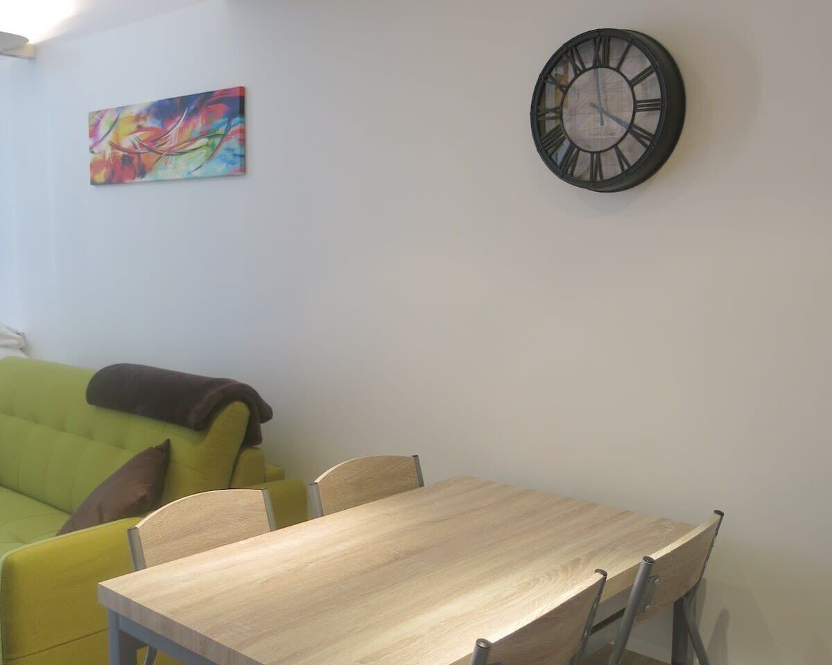 A room with a green sofa, a wooden dining table, and a wall clock.