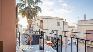 Un balcon avec une table dressée pour deux personnes, agrémentée d’une bouteille de vin et de verres, de bols remplis de nourriture, et offrant une vue sur le paysage urbain.