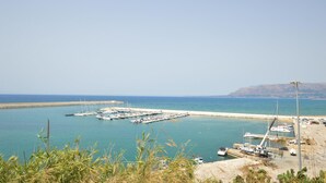 Un port de plaisance situé sur la côte, avec des bateaux amarrés, une jetée s’avançant dans l’eau et une vue dégagée sur la mer et les collines lointaines.