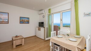 Ein Zimmer mit Esstisch für zwei Personen, einem kleinen Schreibtisch mit Stuhl, einem Fernseher und einem Fenster mit Meerblick.