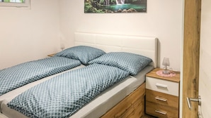 A bed with a white headboard and two pillows, a wooden nightstand with drawers, and a small table with a lamp.