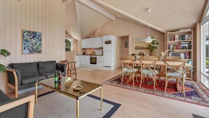 Amplia sala de estar con un sofá, una mesa de centro y un comedor. Se ve una cocina con armarios blancos y una zona de comedor con un tapete rojo estampado.