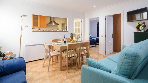Een moderne woonkamer met een blauwe bank, een eettafel met stoelen, en een keuken met een magnetron en een fornuis.