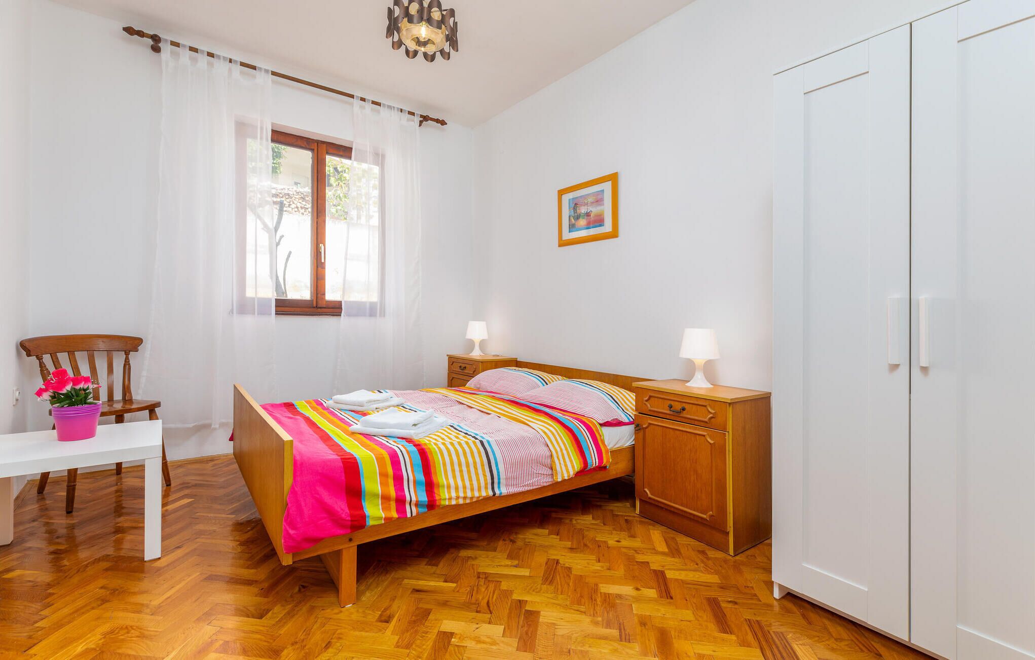 A bedroom with a wooden bed, two bedside tables with lamps, a chair, a small table with a potted plant, a window with sheer curtains, and a door.