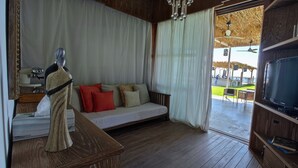 A cozy room with a wooden floor, a sofa with cushions, a television on a wooden stand, and a large window with curtains. - LOST AT SEA (Damour)