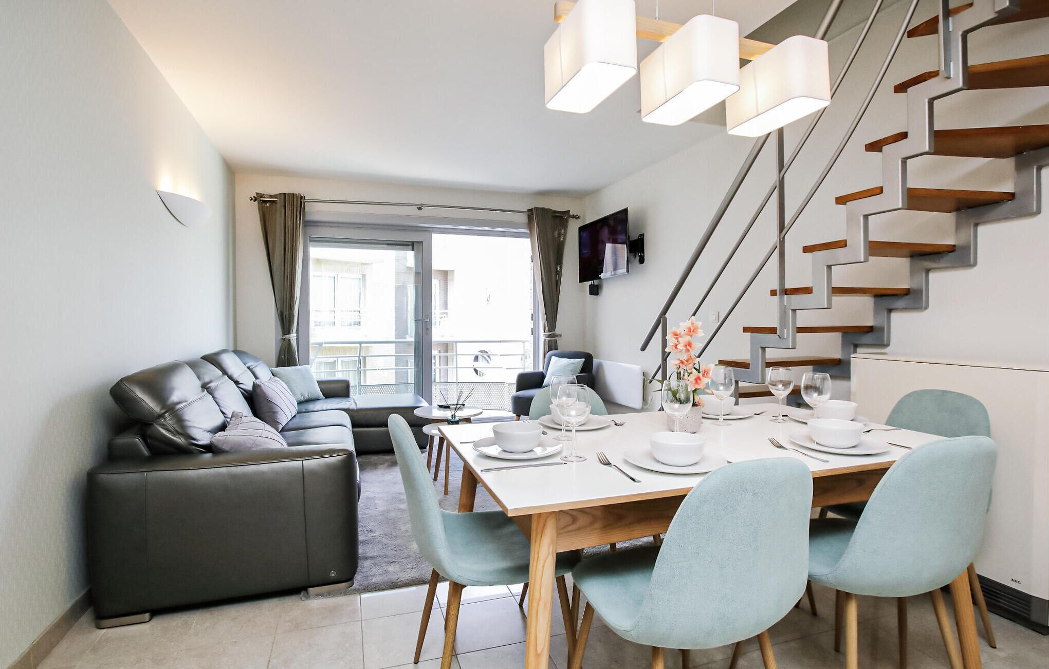 Et moderne spiseområde med et træbord dækket til fire personer, omgivet af lyseblå stole. En sort lædersofa står op ad væggen, og en trappe med trætrapper er synlig i baggrunden.