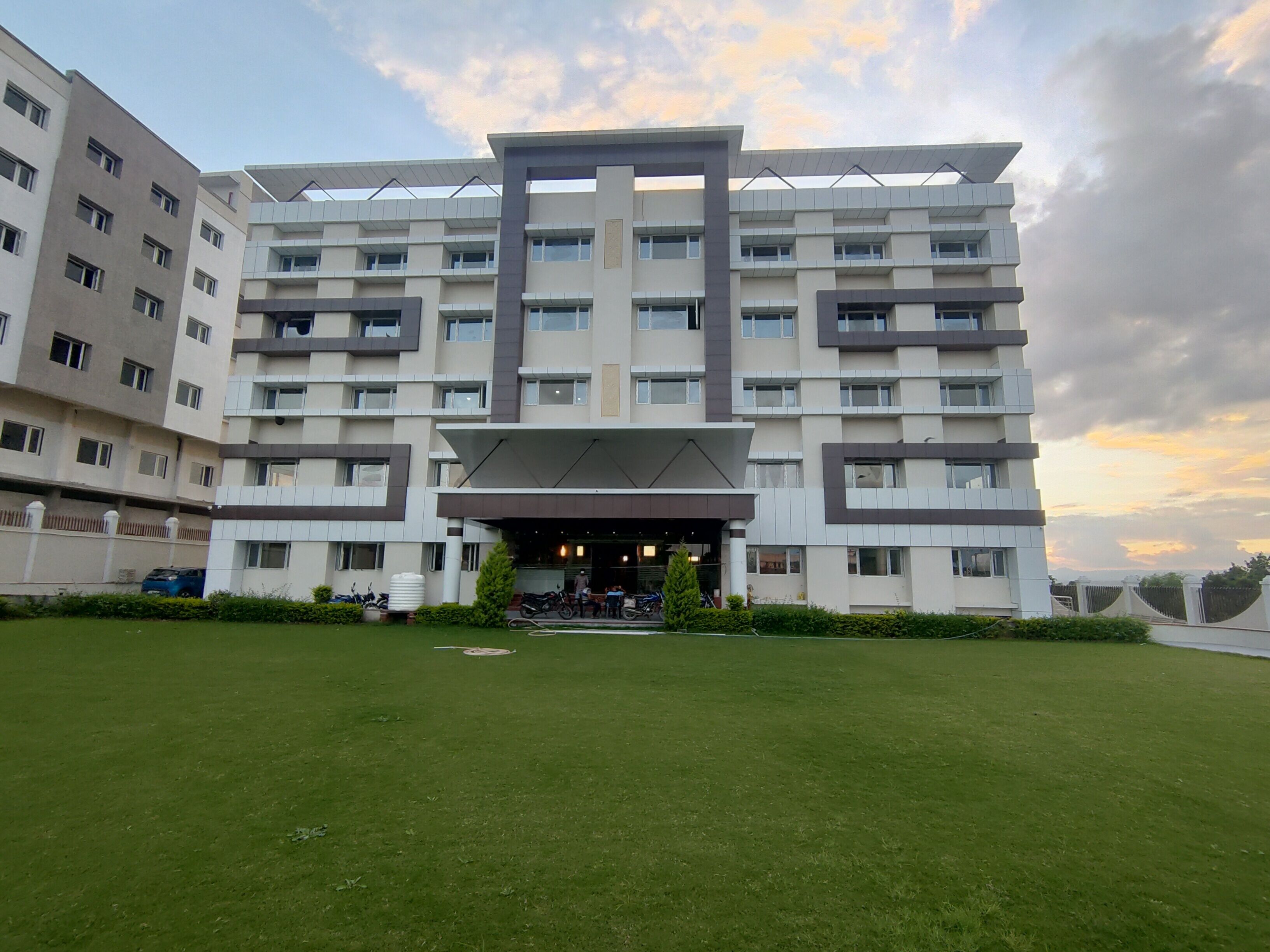 A modern multi-story hotel with a central entrance, surrounded by a well-maintained lawn and a parking area.
