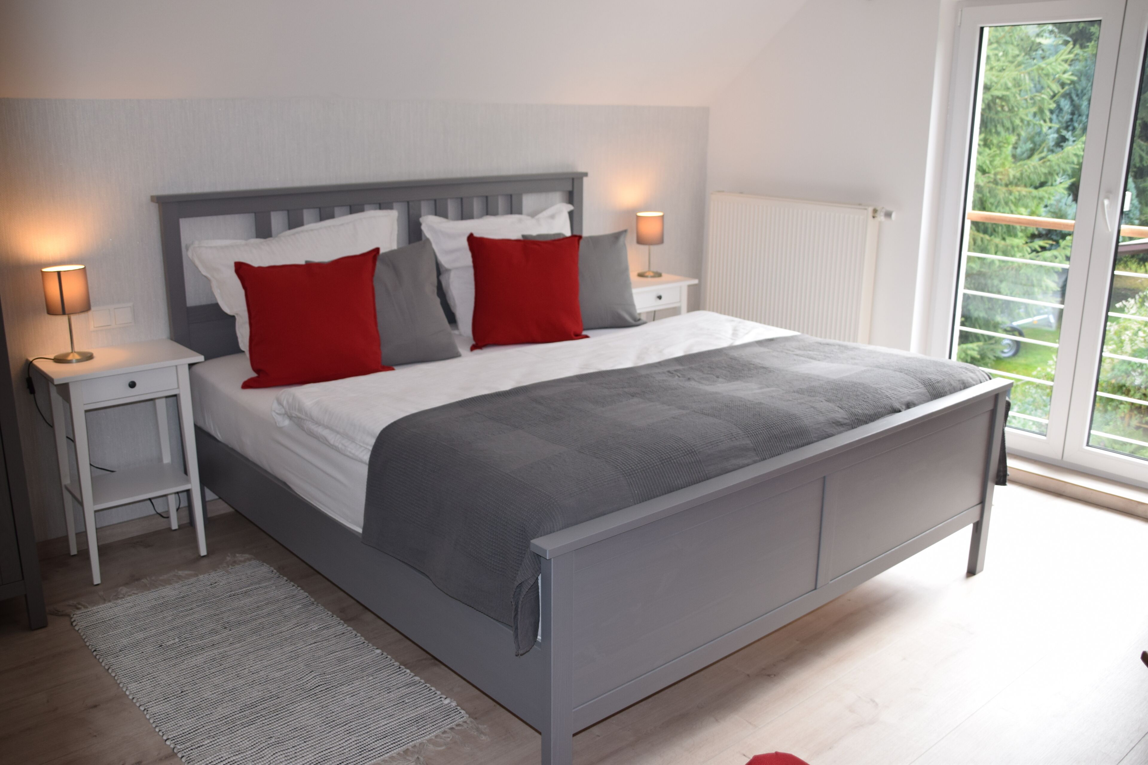 Un dormitorio moderno con una cama gris, almohadas blancas y rojas, un cabecero gris, una mesita de noche blanca con una lámpara, y vista a la vegetación a través de una puerta corrediza de vidrio.
