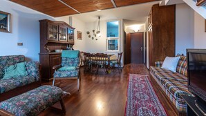 Un salon chaleureux avec un plafond en bois, un coin repas avec des chaises et une table, et un canapé agrémenté d’un plaid à motifs.