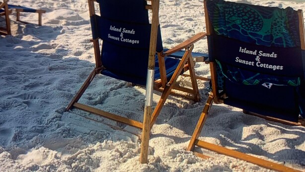 Deux chaises de plage avec un parasol bleu, portant l’inscription « Island Sands & Sunset Cottages », sur une plage de sable, avec des vagues de l’océan en arrière-plan.
