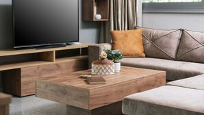 A modern living room with a flat-screen TV on a wooden stand, a coffee table with a book and decorative items, a sofa with cushions, and a window with curtains.