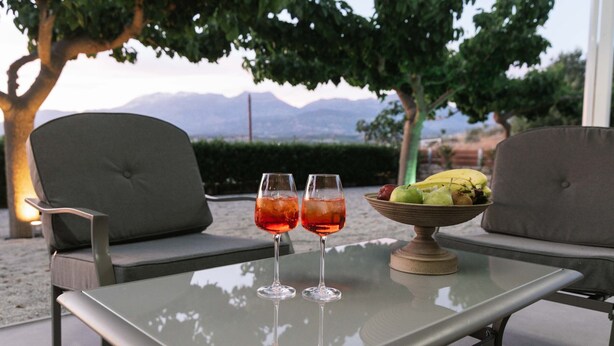 A table with two glasses of red drink, a bowl of fruit, and two chairs with a mountain view in the background.