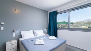 A neatly made bed with white linens and two folded towels, a bedside table with a book, and a window with a view of a green field and hills.