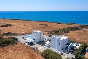 Um edifício moderno branco com piscina, situado numa região seca e montanhosa com vista para o mar.