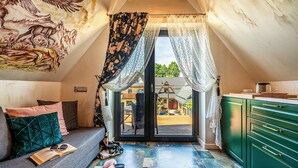 Ett mysigt sovrum på vinden med ett stort fönster, en soffa med kuddar, ett litet läsbord och en grön byrå.