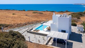 Un edificio moderno con piscina en la azotea, rodeado de hierba seca y árboles, con vistas a un mar azul y despejado.