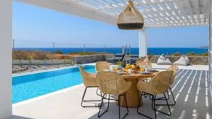 Un área de comedor junto a la piscina con sillas de mimbre, una mesa de madera y una lámpara colgante.