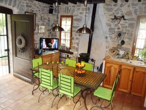 Una cocina rústica con pared de piedra, armarios de madera, una mesa de comedor con sillas verdes y un televisor sobre un mueble de madera.