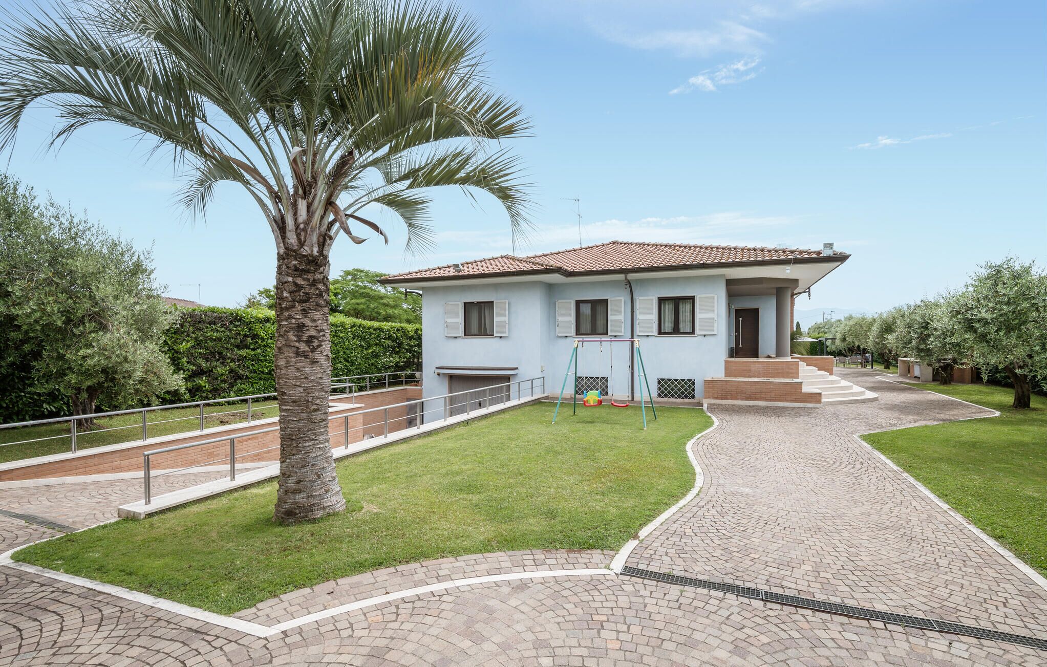 Una casa a un piano con tetto in tegole, vialetto lastricato e un piccolo giardino con una palma.