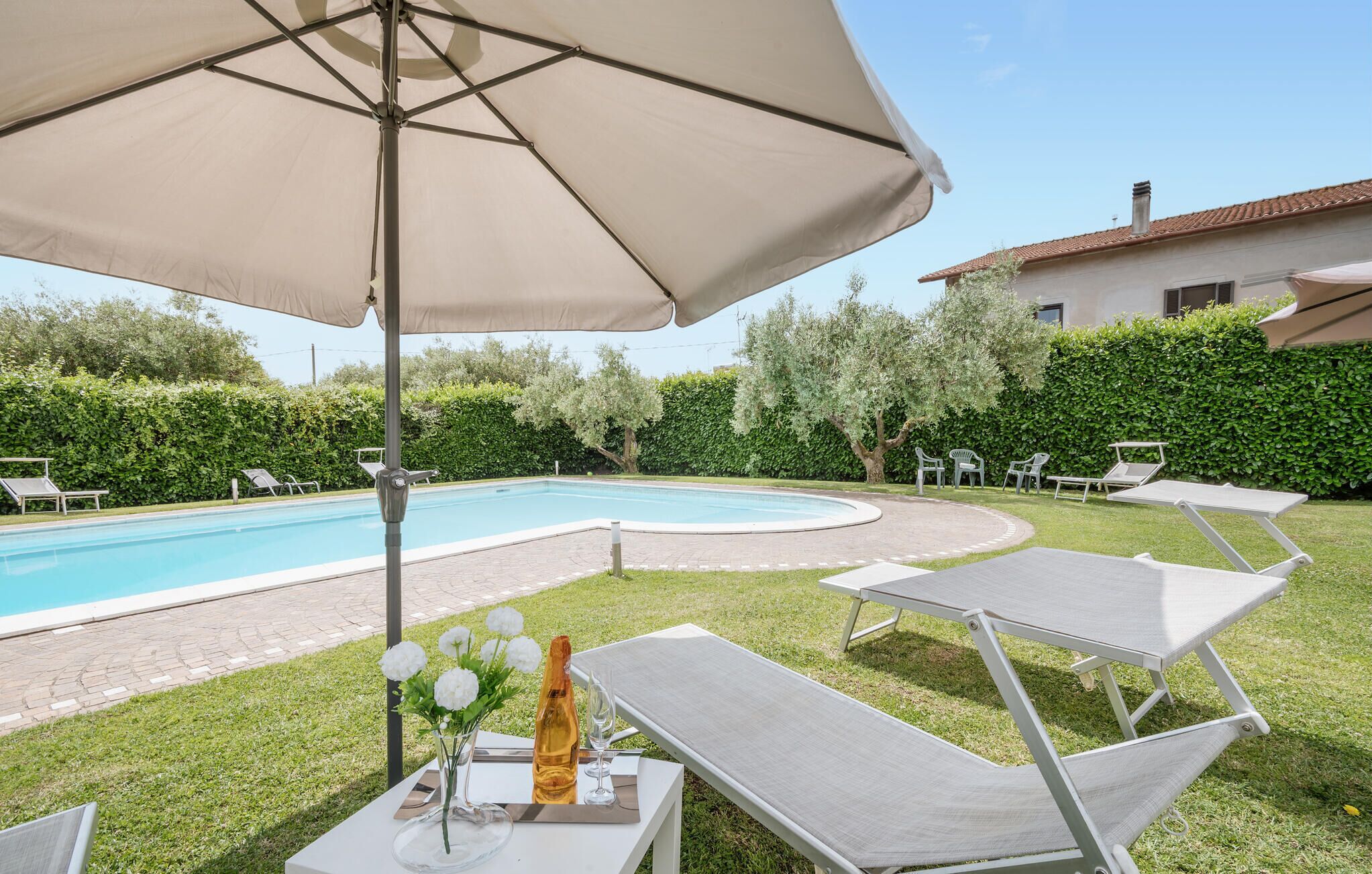 Un espace aménagé au bord de la piscine, avec des chaises longues, une table ornée d’un vase de fleurs et d’une bouteille, et un grand parasol.