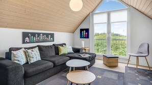 Una sala moderna con un sofá seccional gris oscuro, una mesa de centro redonda, un reposapiés de mimbre y un sillón gris. Hay dos cuadros en la pared, una ventana con vistas a zonas verdes y un techo de madera.