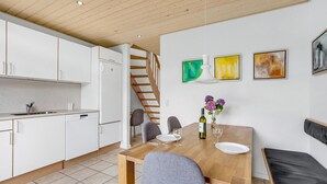 Una cocina moderna con armarios blancos, lavavajillas integrado y un refrigerador. Hay un comedor con mesa de madera, sillas grises y un banco. La habitación tiene techo de madera y una escalera que conduce a otro nivel.