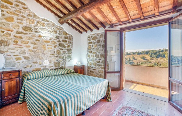 Quarto com parede de pedra, teto de madeira, cama com roupa de cama listrada, mesa de cabeceira, abajur e varanda com vista para o campo.