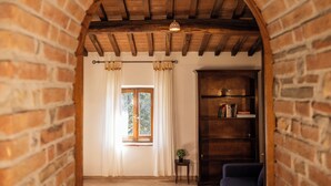 Una habitación con pared de ladrillo, vigas de madera en el techo, una estantería, una ventana con cortinas y una pequeña mesa con una planta.