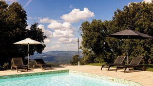 Piscina con sillas de descanso y sombrillas, rodeada de árboles y vistas a las montañas.