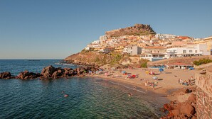Un pueblo costero con una imponente fortaleza en la cima de una colina, playa de arena y mar azul cristalino.