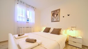 Ein ordentlich eingerichtetes Schlafzimmer mit einem Bett, zwei Nachttischen mit Lampen, einem Heizkörper und einem Fenster mit transparenten Vorhängen.