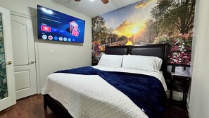 A bedroom with a bed, a TV mounted on the wall, and a mural of a sunset scene. - GaMe RoOm, Sleep 12, Colorful Back Yard, Beach(A) (Long Beach)