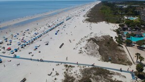 Una playa con numerosos sombrillas y sillas, un sendero y una piscina con un edificio cercano.