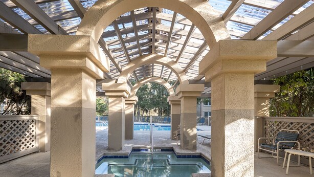 Una piscina interior con techo de cristal, rodeada de pilares color beige, y una zona junto a la piscina con una silla y una mesa.