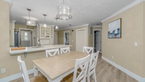 Un comedor con mesa de madera y sillas blancas, una cocina con armarios blancos e iluminación colgante.