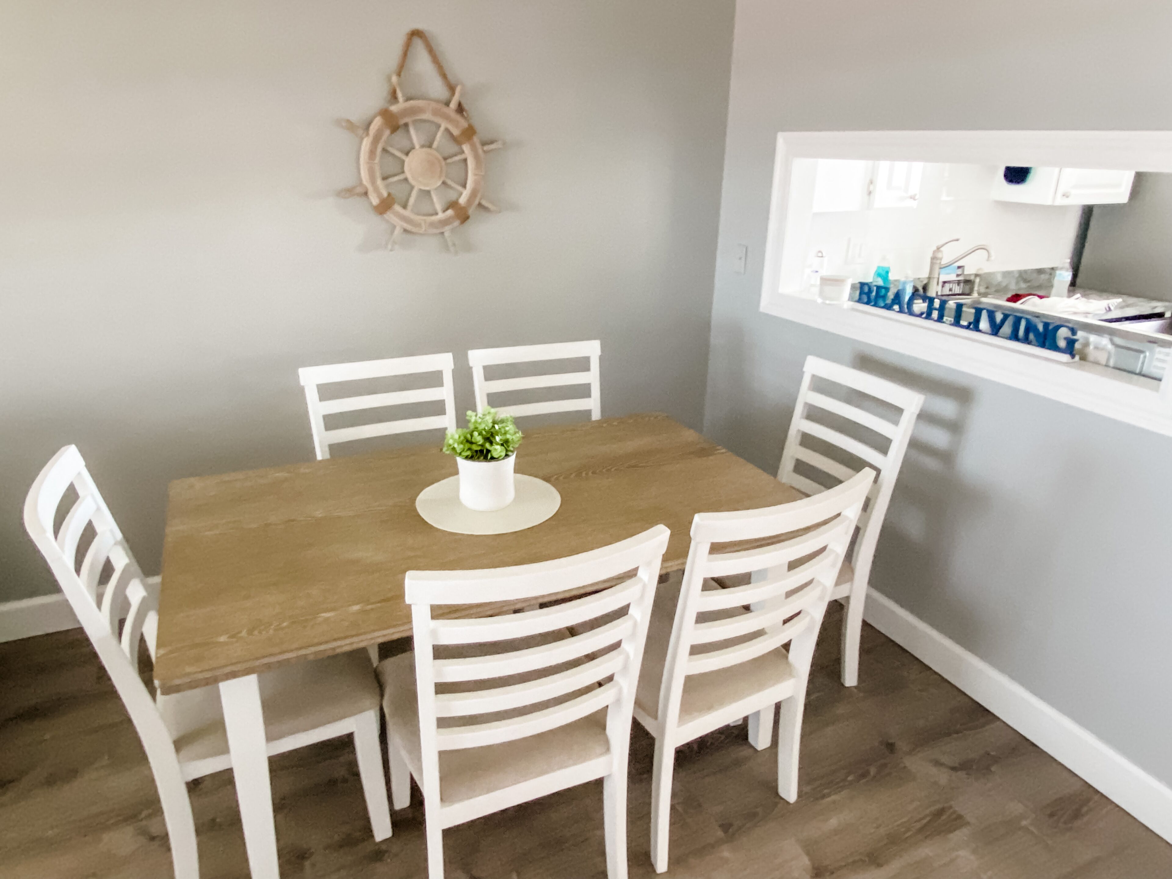 Un comedor con una mesa de madera y sillas blancas, una planta pequeña en maceta y un adorno de timón de barco colgado en la pared.