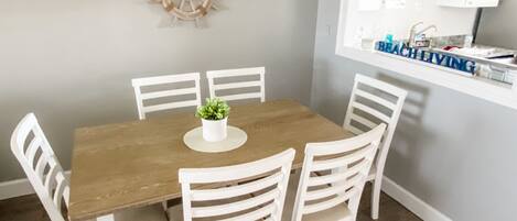 Un comedor con una mesa de madera y sillas blancas, una planta pequeña en maceta y un adorno de timón de barco colgado en la pared.